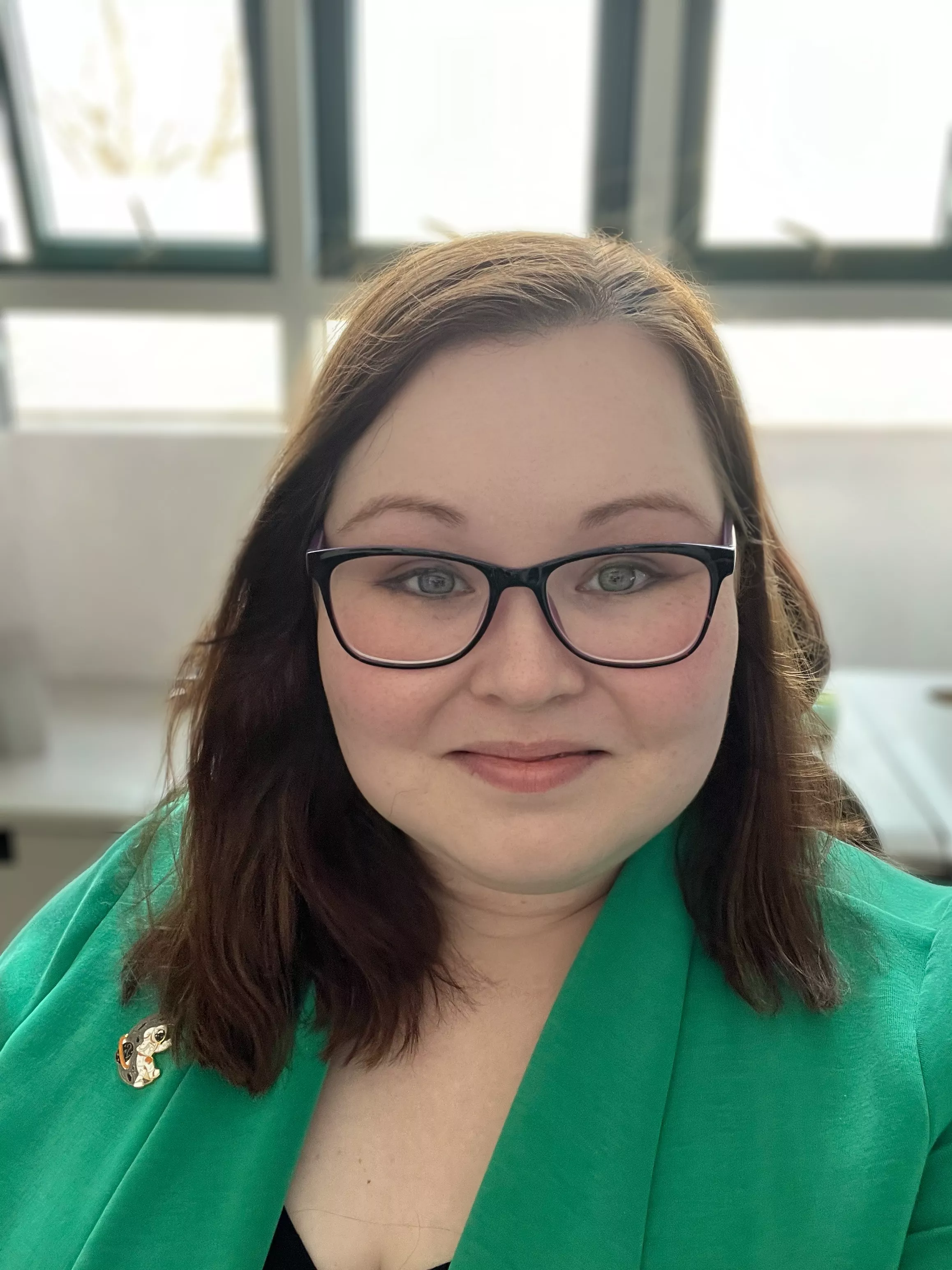 A smiling woman with fair skin, long brown hair, and black glasses wears a green blazer with a decorative pin. She is indoors with bright natural light coming through large windows behind her.